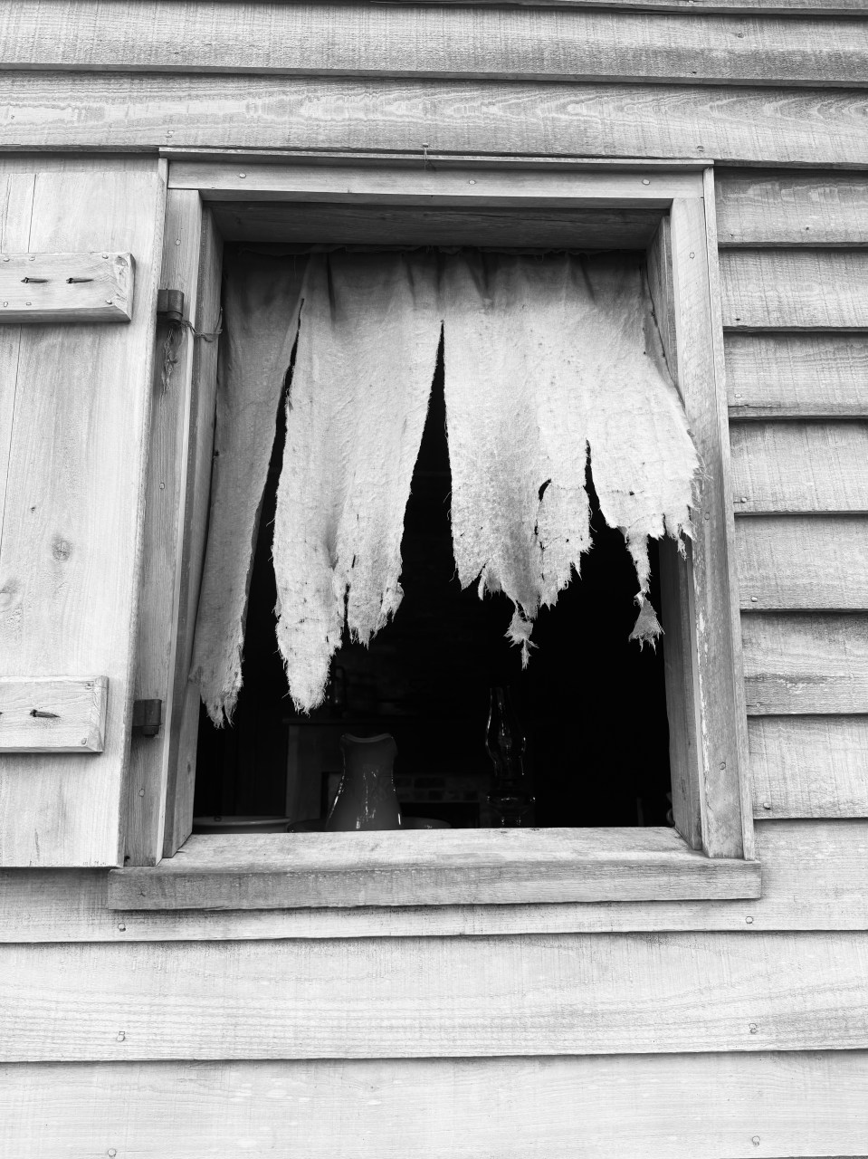 Old wooden window with torn curtain, showing a jug and a bottle inside.