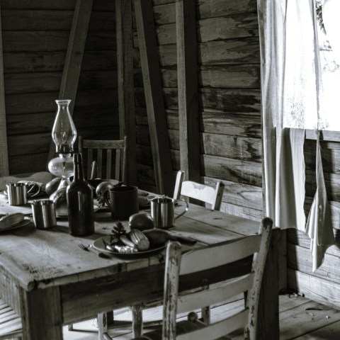 Rustic wooden dining table with chairs, oil lamp, and plates in a dimly lit cabin.